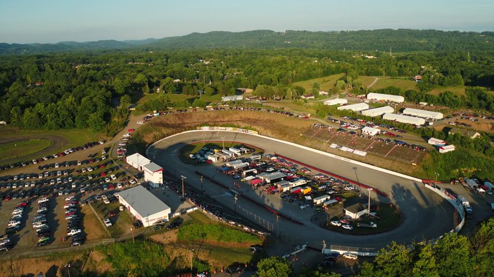 An aerial view of a local race track on a bustling event day, with packed parking areas and a vibrant gathering of spectators and racers
