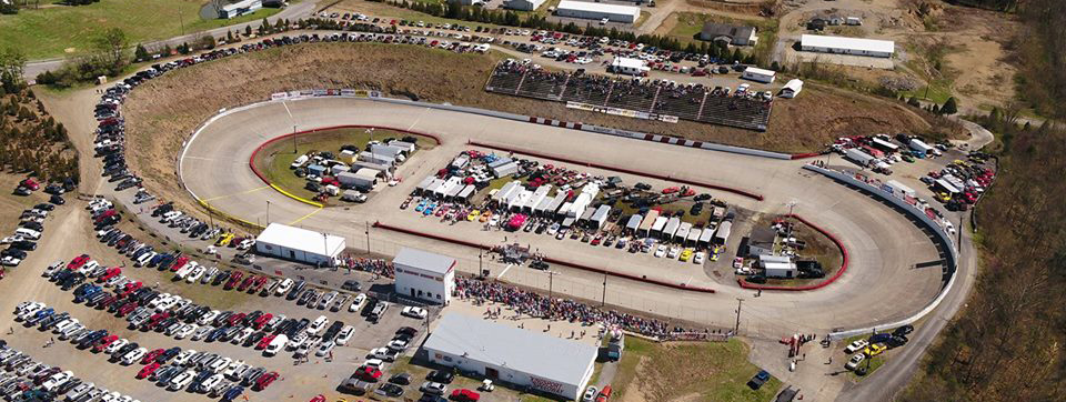 An aerial view of a bustling race track on a sunny day, showcasing a packed parking lot and spectators gathered for an event