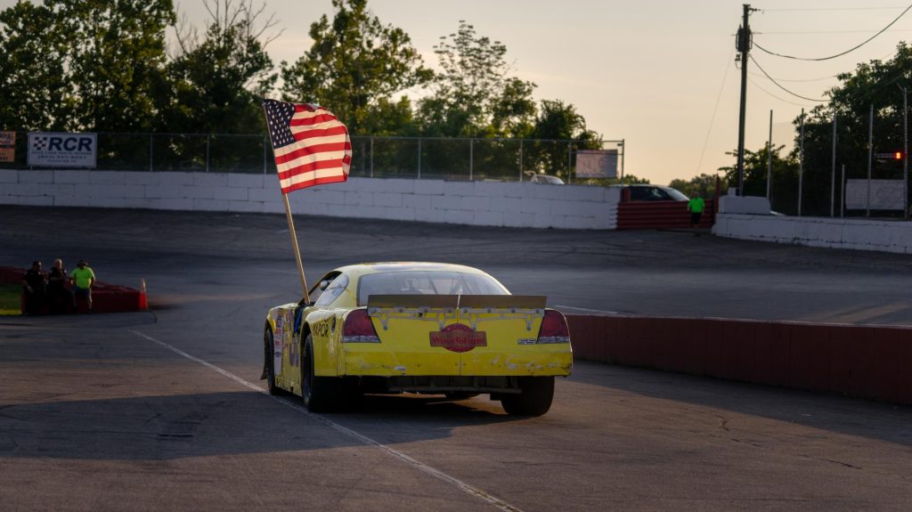 A race car sporting a weathered yellow paint job proudly carrying the american flag as it cruises along a local racetrack at dusk.