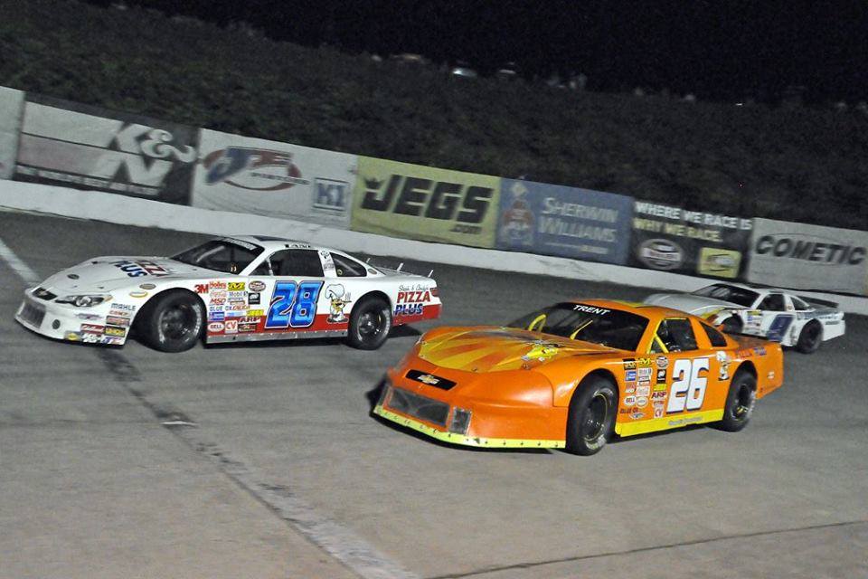 Two race cars fiercely compete on the track, the orange number 26 car taking a tight inside line as it challenges the white number 26 for the lead under the night lights.