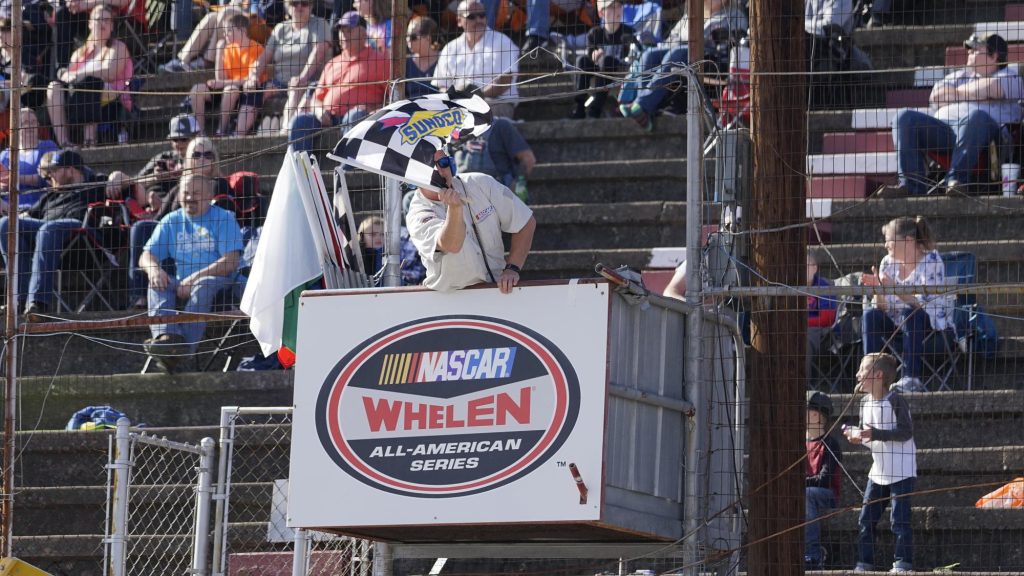 An official at a nascar whelen all-american series race enthusiastically waves the checkered flag, signaling the end of a race, while spectators in the stands look on during a sunny day at the track