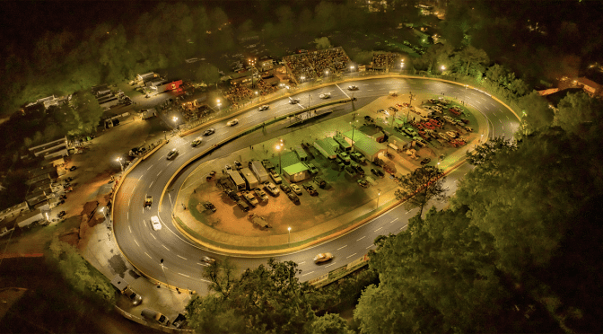 Aerial night view of a brightly lit oval race track surrounded by trees, with cars racing and parked vehicles, including a bustling pit stop area