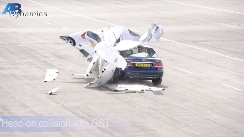 A blue sedan has collided head-on with a large white crash test barrier simulating an accident. debris is scattered around the impact zone on a testing track
