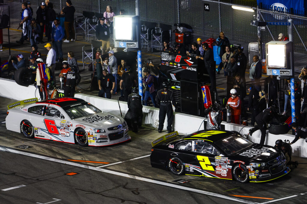 Two race cars, number 6 and 22, speeding past a crowded pit stop area during an evening race at a lit-up track