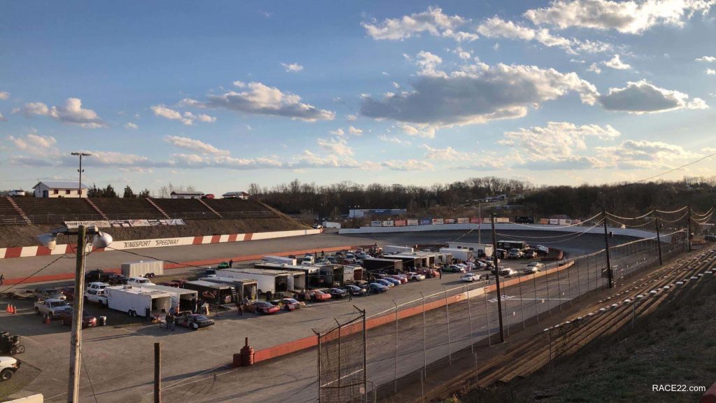 Sunset view of a local race track with cars and trailers gathered in the pit area, surrounded by empty spectator stands and a clear sky