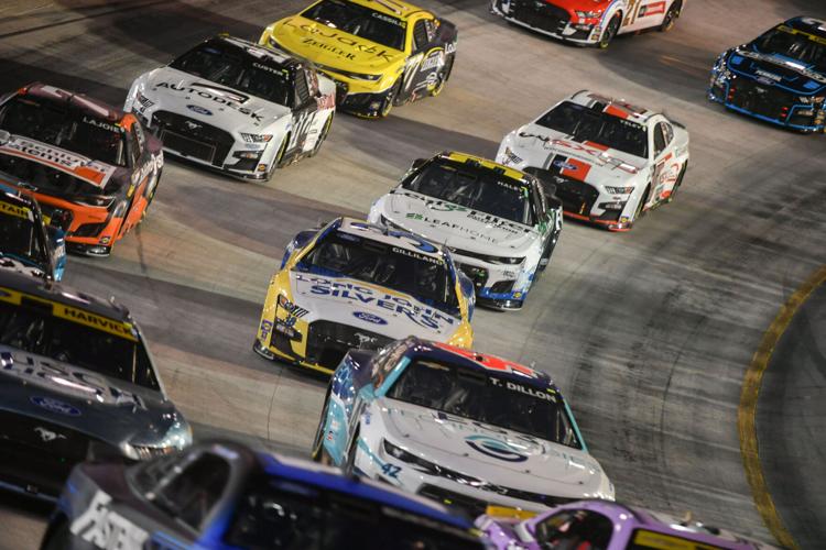 Nascar race cars tightly grouped on a track, showcasing a mix of vibrant colors and sponsor logos, under bright stadium lights