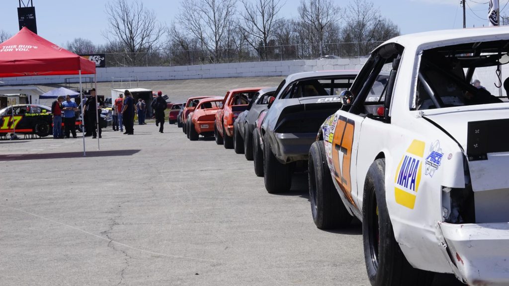 A lineup of race cars parked in the pit area on a sunny day, with people milling about in the background