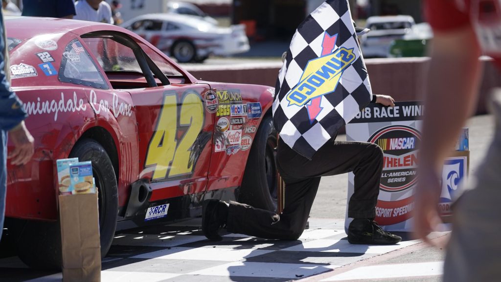 A moment of triumph: a race car emblazoned with the number 47 is welcomed back to the pit after a victorious run, with a crew member crouching by its side, proudly displaying the checkered flag - the classic symbol of a racing win