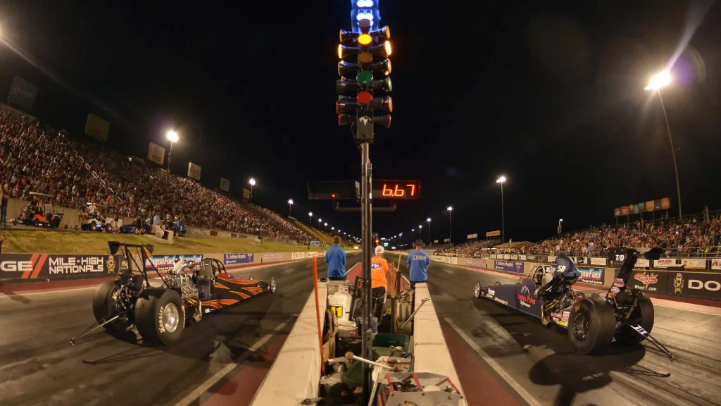 Two drag racing cars at a start line with the green light on, viewed from the perspective of the start line crew, with a crowded audience on both sides of the track at night